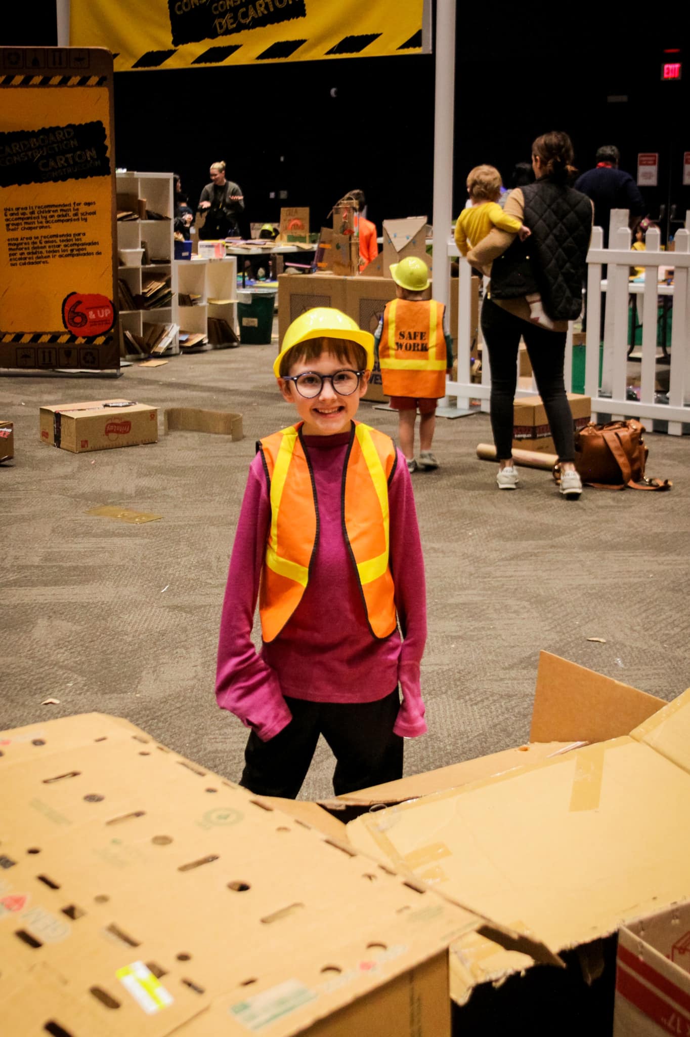 Young boy wearing a safety hat and vest standing in front of a cardboard sculpture. 